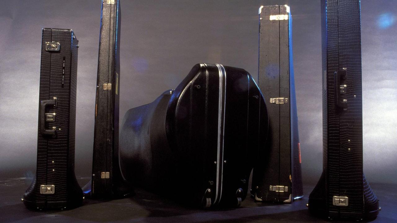 Fünft schwarze, geschlossene Lederkoffer vor grau-silbrigen Hintergrunde, denen man durch ihre geschwungenen Formen ansehen kann, dass darin Blasinstrumente aufgewahrt werden.