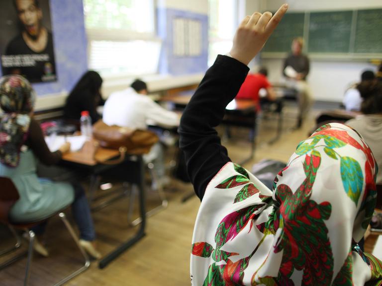 Eine Klasse beim Schulunterricht, die Tische stehen in Zweierreihen, im Hintergrund der Lehrer vor der Tafel, im Vordergrund ein Mädchen mit Kopftuch, das sich meldet.