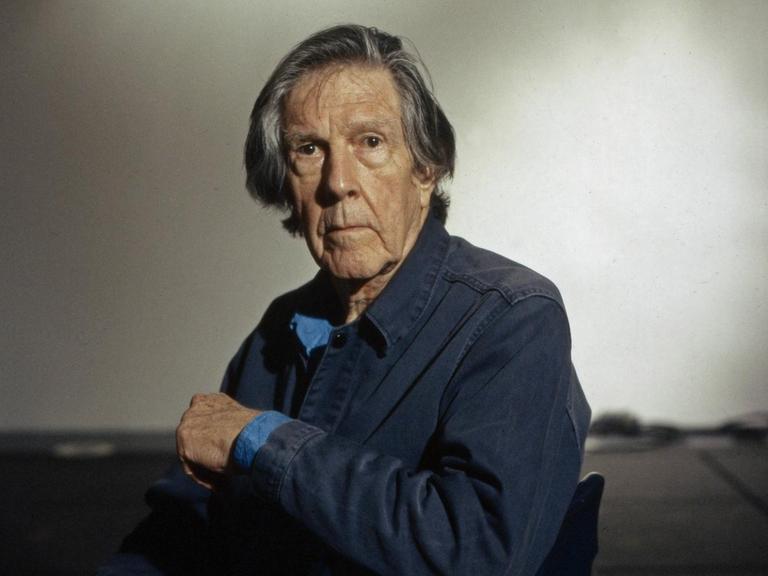 John Cage sitzt seitlich auf einem Stuhl und blickt in die Kamera