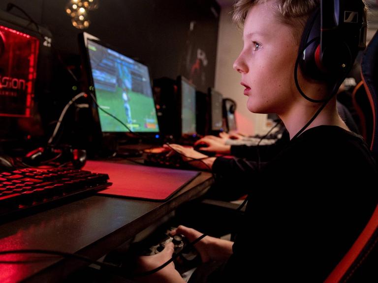 Ein Junge sitzt vor einem Computermonitor und spielt das Spiel "Fortnite". Sein Blick ist gefesselt und in der Hand hält er eine Fernbedienung.