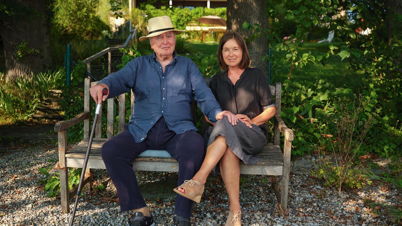 Martin Walser sitzt links von seiner ältesten Tochter Franziska Walser auf einer Bank, er legt seine Hand auf das Knie von ihr. Im Hintergrund Bäume und Pflanzen. Überlingen, 8/2020