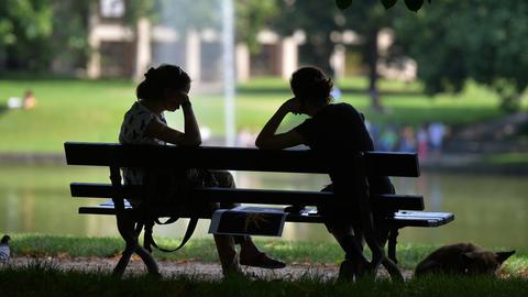 Zwei Frauen sitzen im Schatten auf einer Parkbank, nur ihre Silhouetten sind zu erkennen.