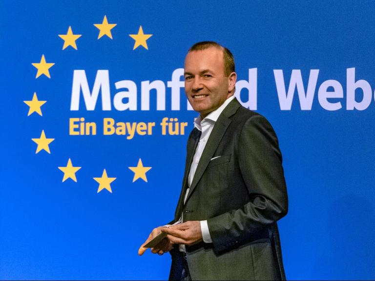 Weber geht lächelnd an einer blauen Wand mit dem europäischen Sternenkreis vorbei, auf der steht: "Manfred Weber - ein Bayer für Europa".