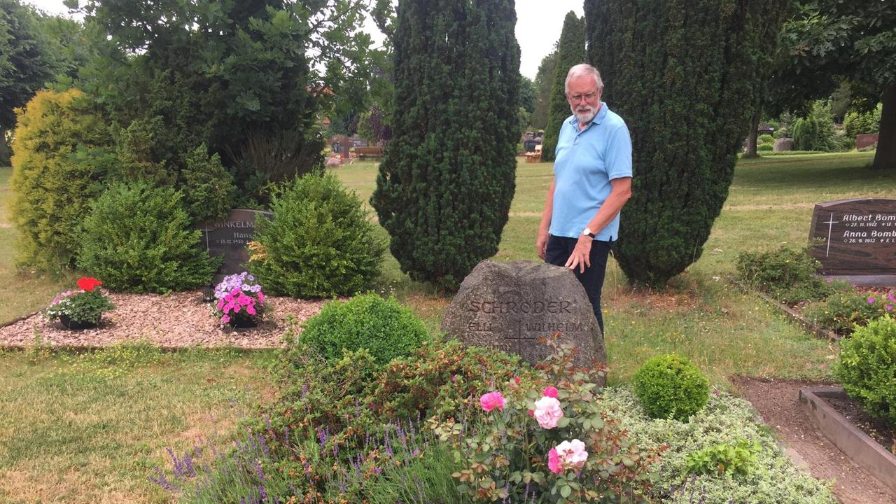Ein grauhaariger Mann mit hellblauem, kurzärmeligen Hemd steht auf einem Friedhof vor einem Grab, das von üppig gewachsenen Pflanzen bedeckt ist. 