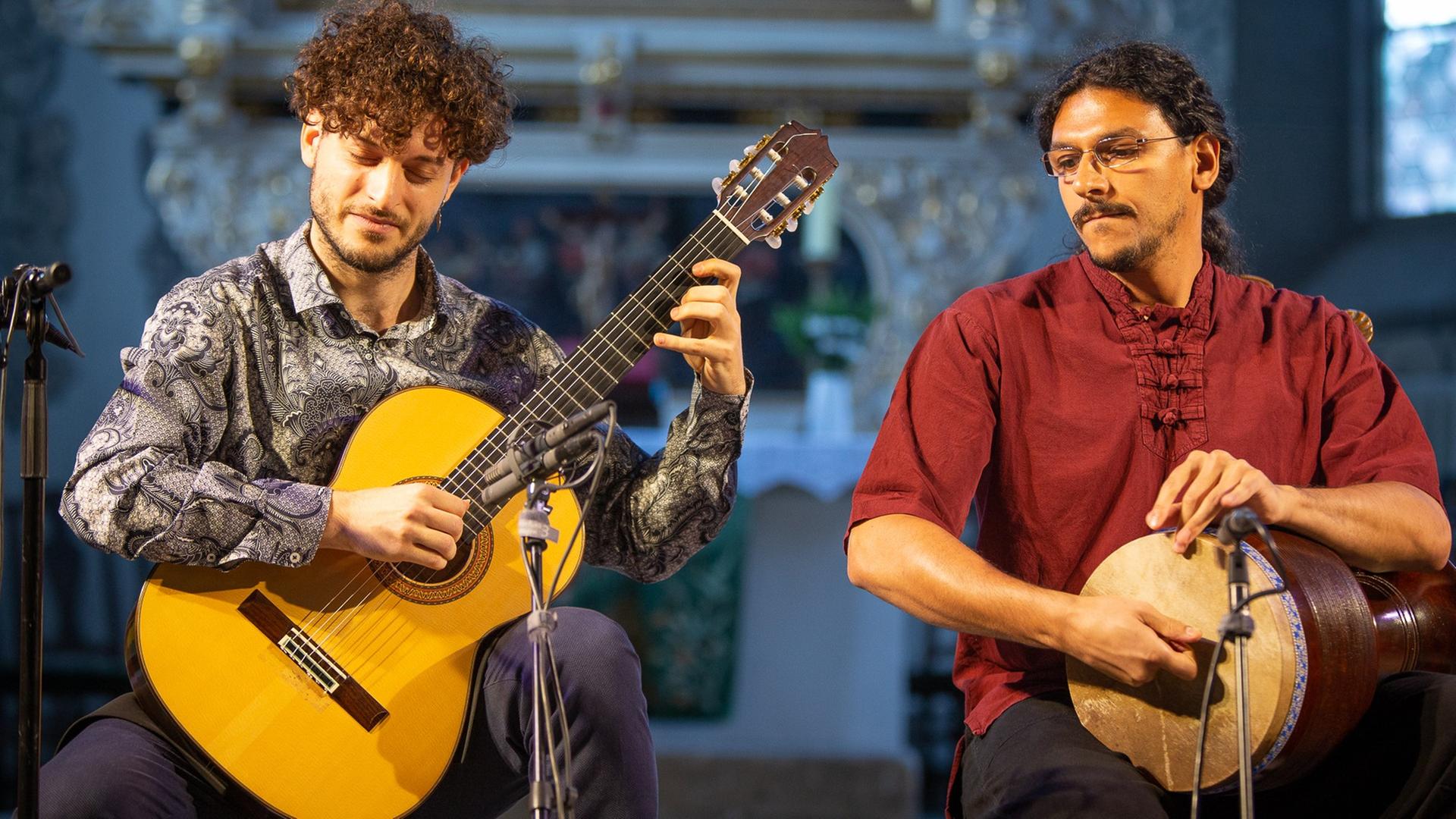 Zwei Männer sitzen auf einer Bühne: Einer spielt Gitarre, der andere trommelt.