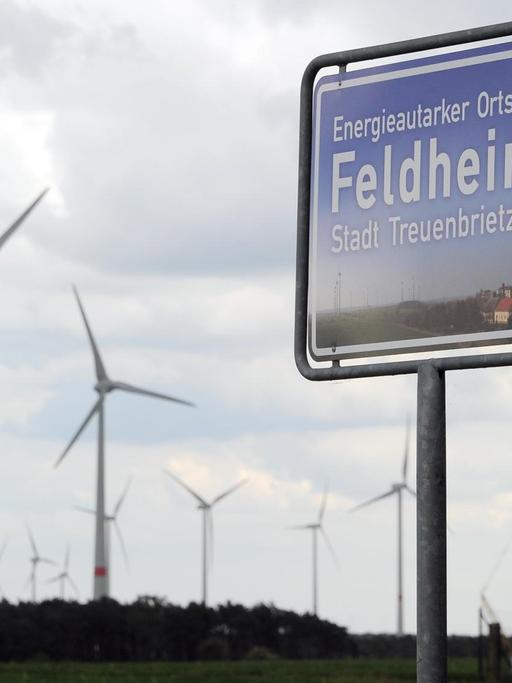 Ein blaues Schild, hinter dem sich Windräder drehen, weist Feldheim (Brandenburg) am 08.05.2014 als energieautarken Ort aus.