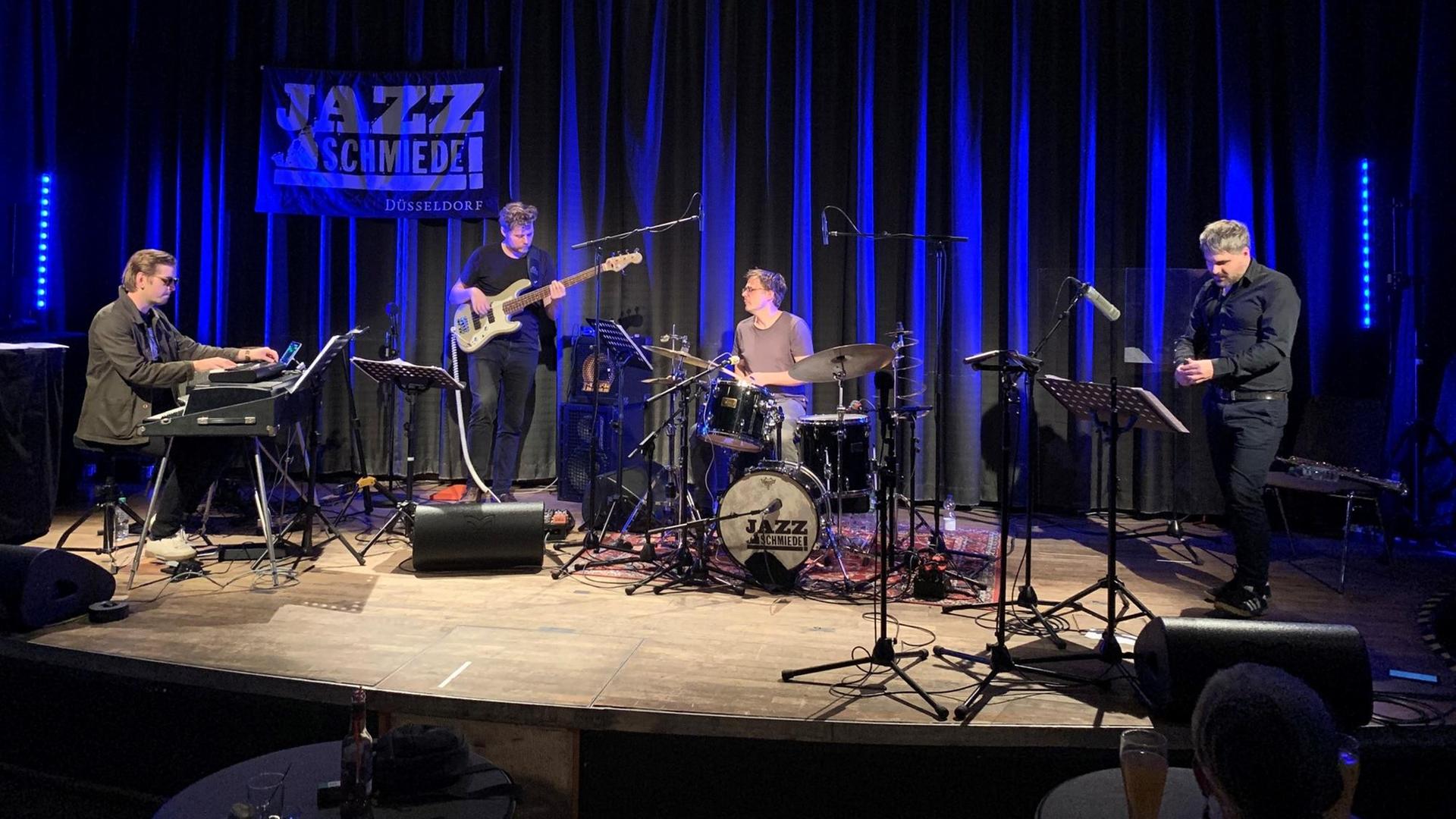 Auf einer Bühne spielen v.l. ein Keyboarder, Bassist, Drummer und Saxofonist zusammen, im Hintergrund ist ein blau angeleuchteter Vorhang und ein Plakat der Jazzschmiede zu sehen.