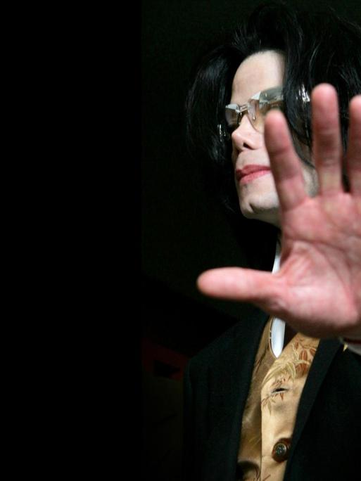 Michael Jackson streckt die Handlfäche der linken Hand Richtung Kamera. Er trägt eine Sonnenbrille. Der Hintergrund ist schwarz.