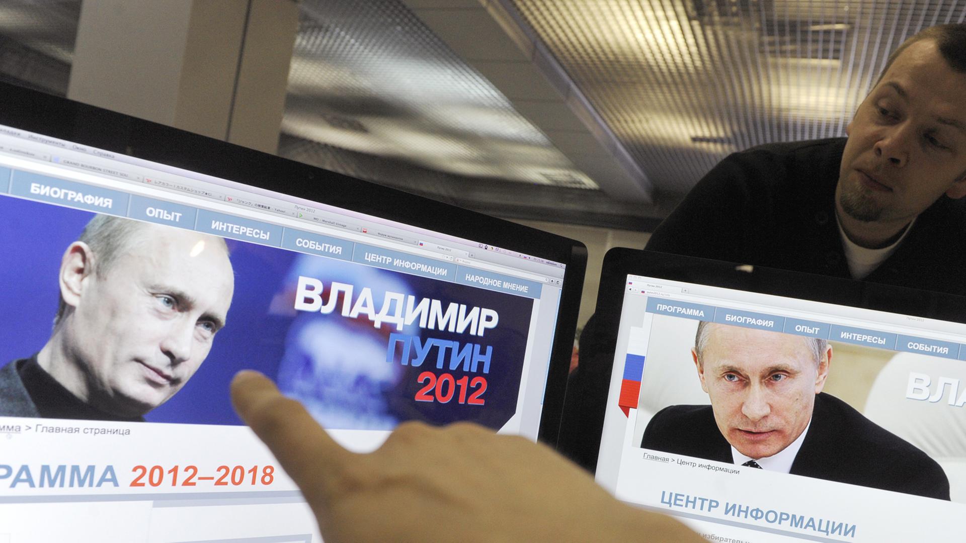 Ein Finger zeigt auf zwei Computerbildschirme, auf denen der russische Präsident Wladimir Putin zu sehen ist.