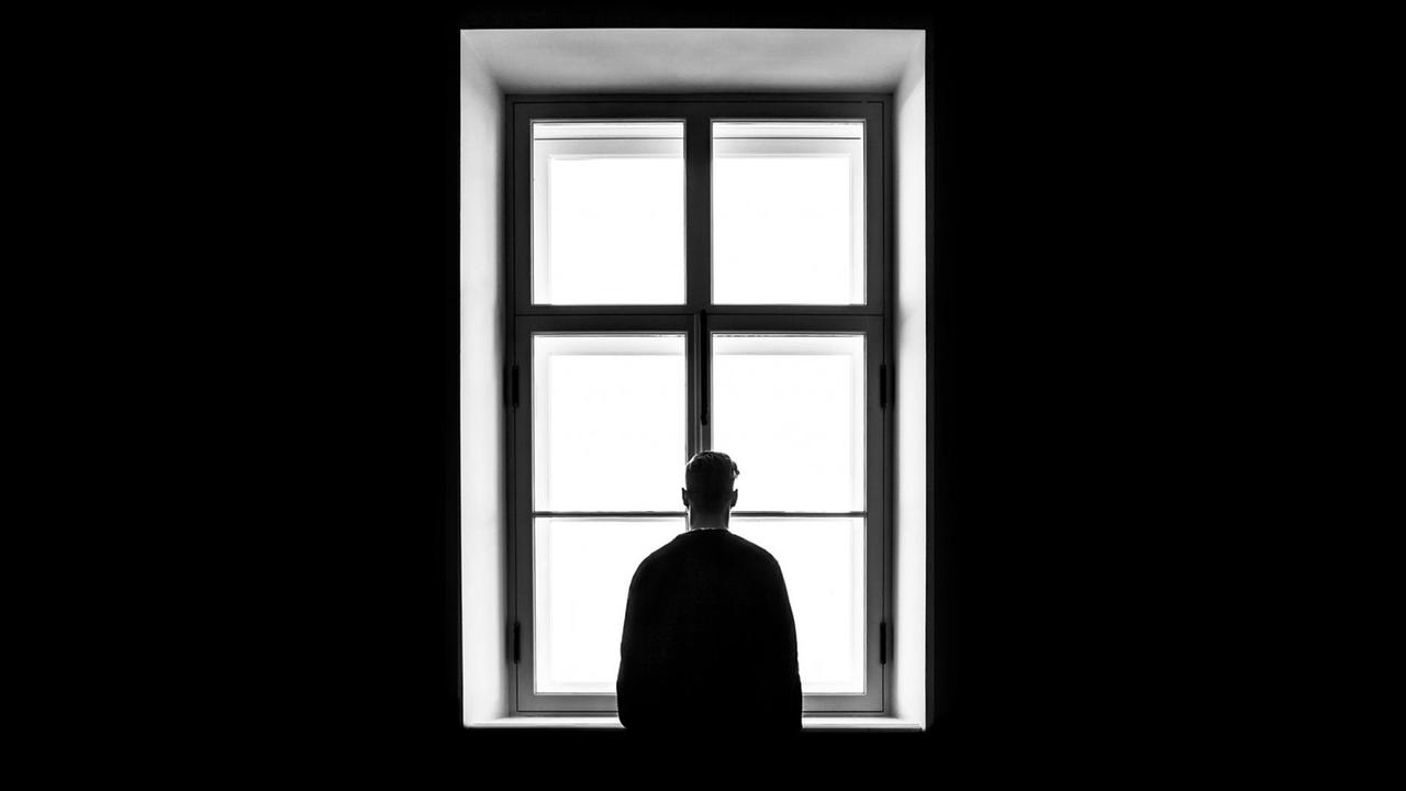 SW-Foto von einem Mann, der dem Betrachter den Rücken zukehrt und vor einem Fenster steht.