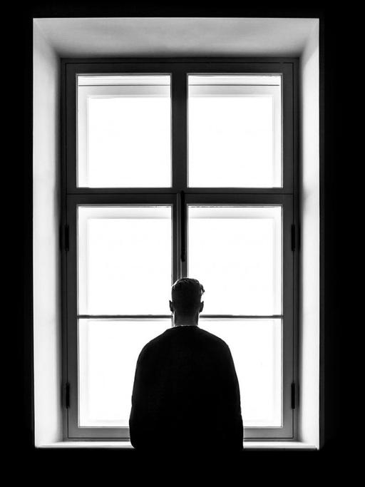 SW-Foto von einem Mann, der dem Betrachter den Rücken zukehrt und vor einem Fenster steht.