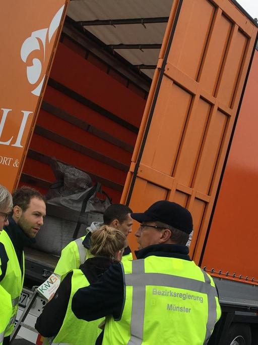 Das Bild zeigt einen orangefarbenen LKW auf einem Autobahnparkplatz zur Abfallkontrolle auf der A2 Im Vordergrund stehen fünf Mitarbeiter der Bezirksregierung Münster.