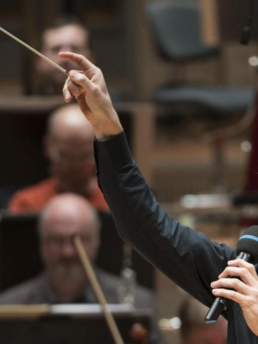 Dirigent Robin Ticciati mit Taktstock und Mikrofon in der Hand moderiert ein Konzert in der Berlliner Philharmonie.