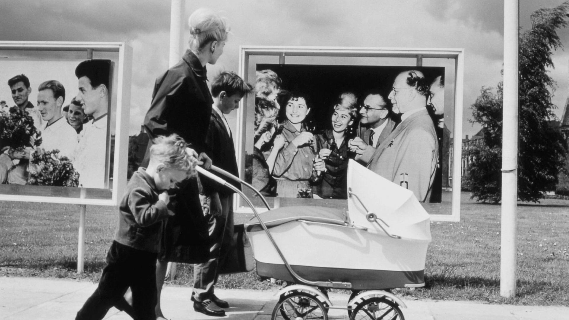 Mutter mit ihren Kindern und einem Kinderwagen, unterwegs in Ostberlin, vor Fotowänden mit SED Propaganda (Fotowand rechts: SED-Generalsekretär Walter Ulbricht u. a.). Foto, 1964. |