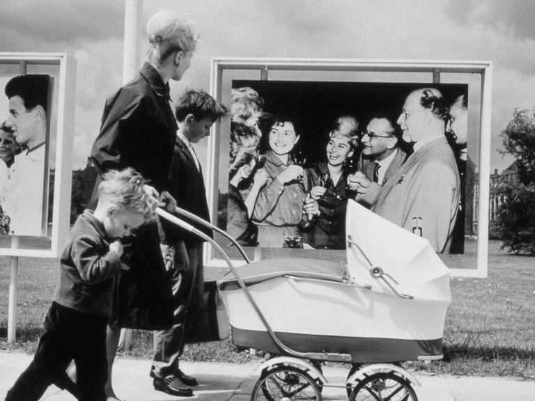 Mutter mit ihren Kindern und einem Kinderwagen, unterwegs in Ostberlin, vor Fotowänden mit SED Propaganda (Fotowand rechts: SED-Generalsekretär Walter Ulbricht u. a.). Foto, 1964. |