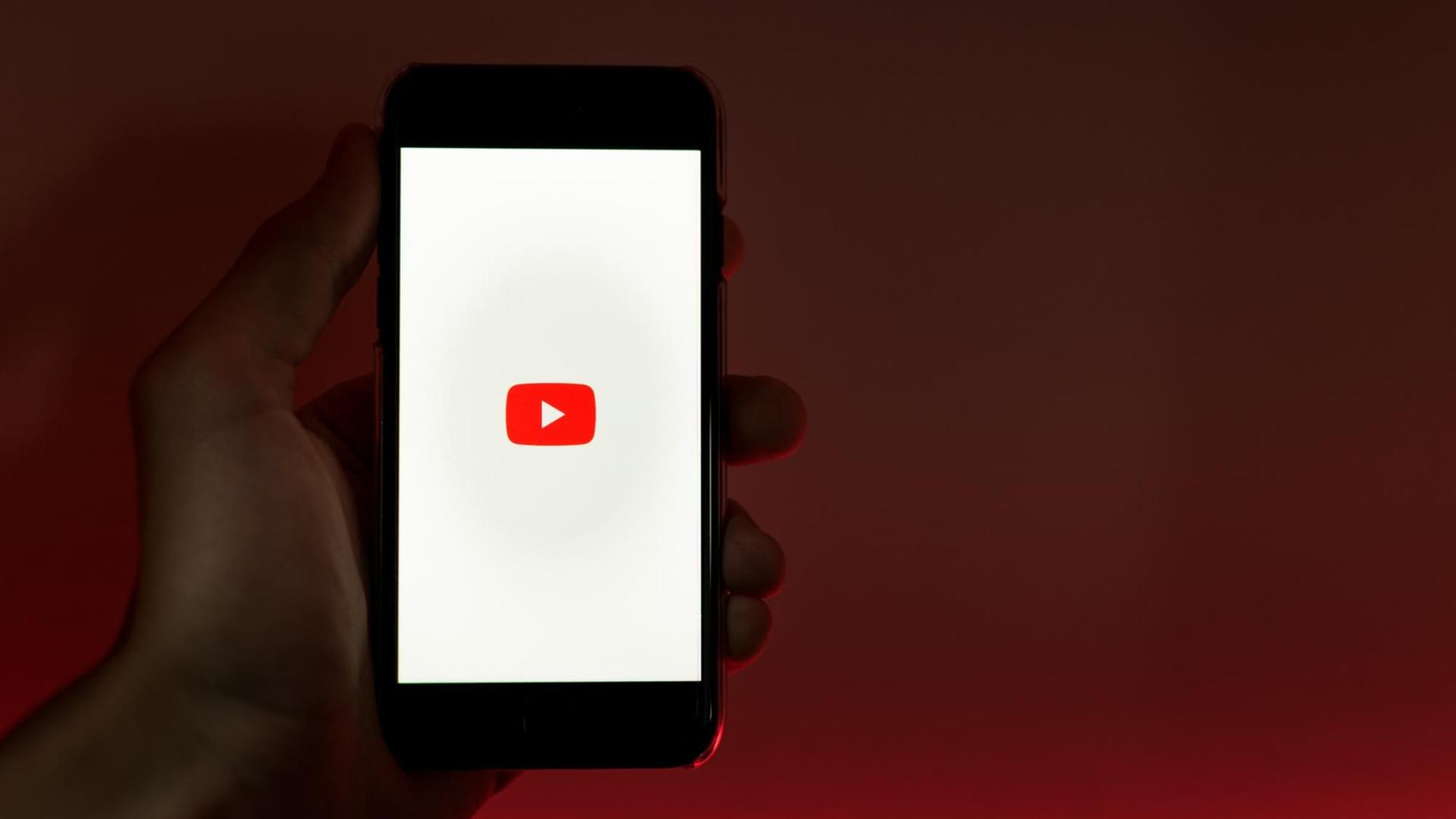 Eine Hand hält ein Smartphone in die Kamera, auf dem das Logo von Youtube abgebildet ist.
