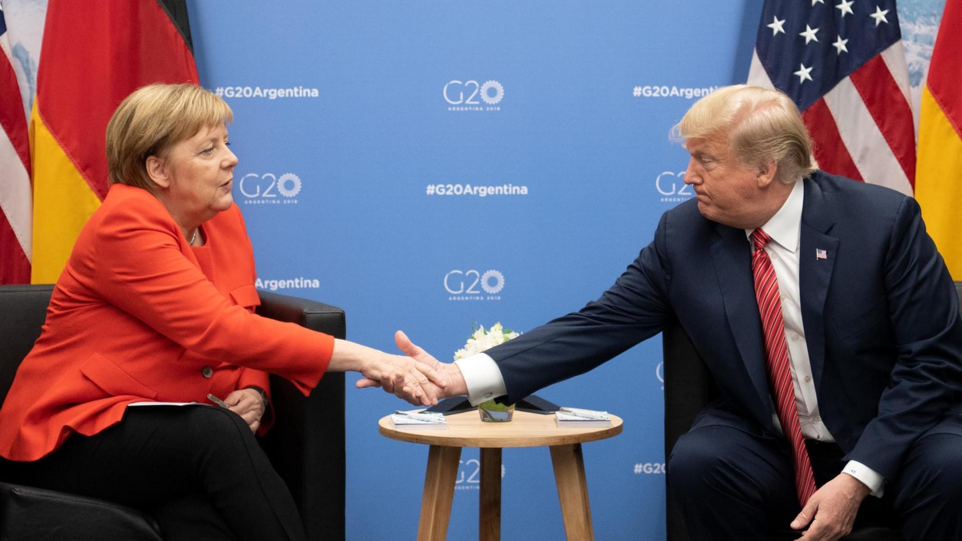 Das Bild zeigt Bundeskanzlerin Angela Merkel (CDU) und Donald Trump, Präsident der Vereinigten Staaten, Sie sitzen nebeneinander an einem Tisch und geben sich die Hand. Im Hinergrund sind die Flaggen Deutschlands und der USA sowie eine blaue Wand mit dem offiziellen Logo des Gipfeltreffens zu sehen.