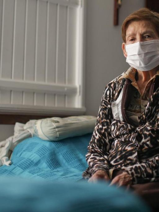 Eine ältere Dame mit Mundschutz sitzt alleine auf einem Bett. Auch in Bogota, Kolumbien werden alle älteren Menschen ab 70 Jahren gebeten, ihr Haus nicht zu verlassen. 18. März 2020