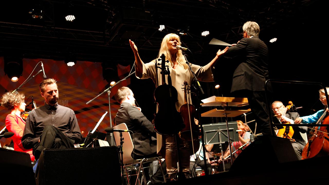 Lena Willemark steht mit ausgebreiteten Armen am Mikrophon vor den Thüringer Symphonikern