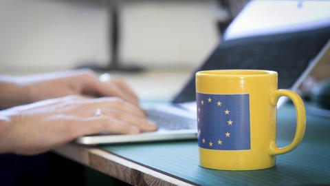 Eine Tasse mit der Flagge Europas Schreibtisch, an dem eine Person am Laptop sitzt.
