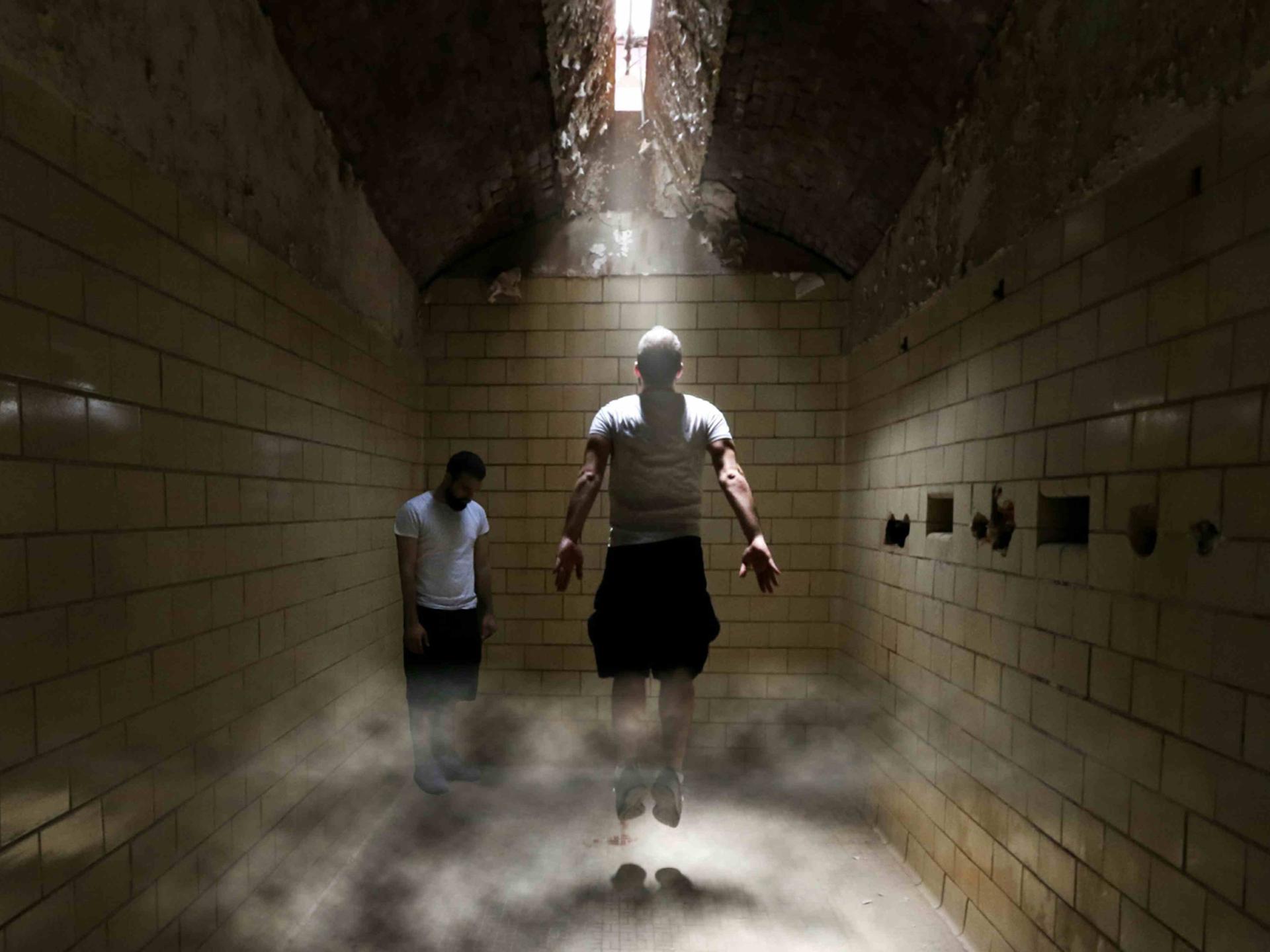 Zwei Menschen stecken in einem unterirdischen Trakt fest, einer von beiden springt nach oben, um näher an eine Luke in der Decke zu kommen, durch die Licht hineindringt.