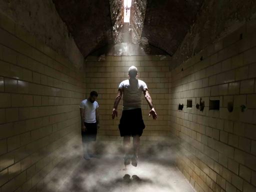 Zwei Menschen stecken in einem unterirdischen Trakt fest, einer von beiden springt nach oben, um näher an eine Luke in der Decke zu kommen, durch die Licht hineindringt.