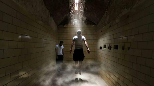 Zwei Menschen stecken in einem unterirdischen Trakt fest, einer von beiden springt nach oben, um näher an eine Luke in der Decke zu kommen, durch die Licht hineindringt.