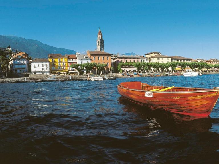 Das Bild zeigt blaues Wasser mit einem roten Boot im Vordergrund, dahinter ist ein Hafen und ein Berg zu sehen, der Monte Verità.