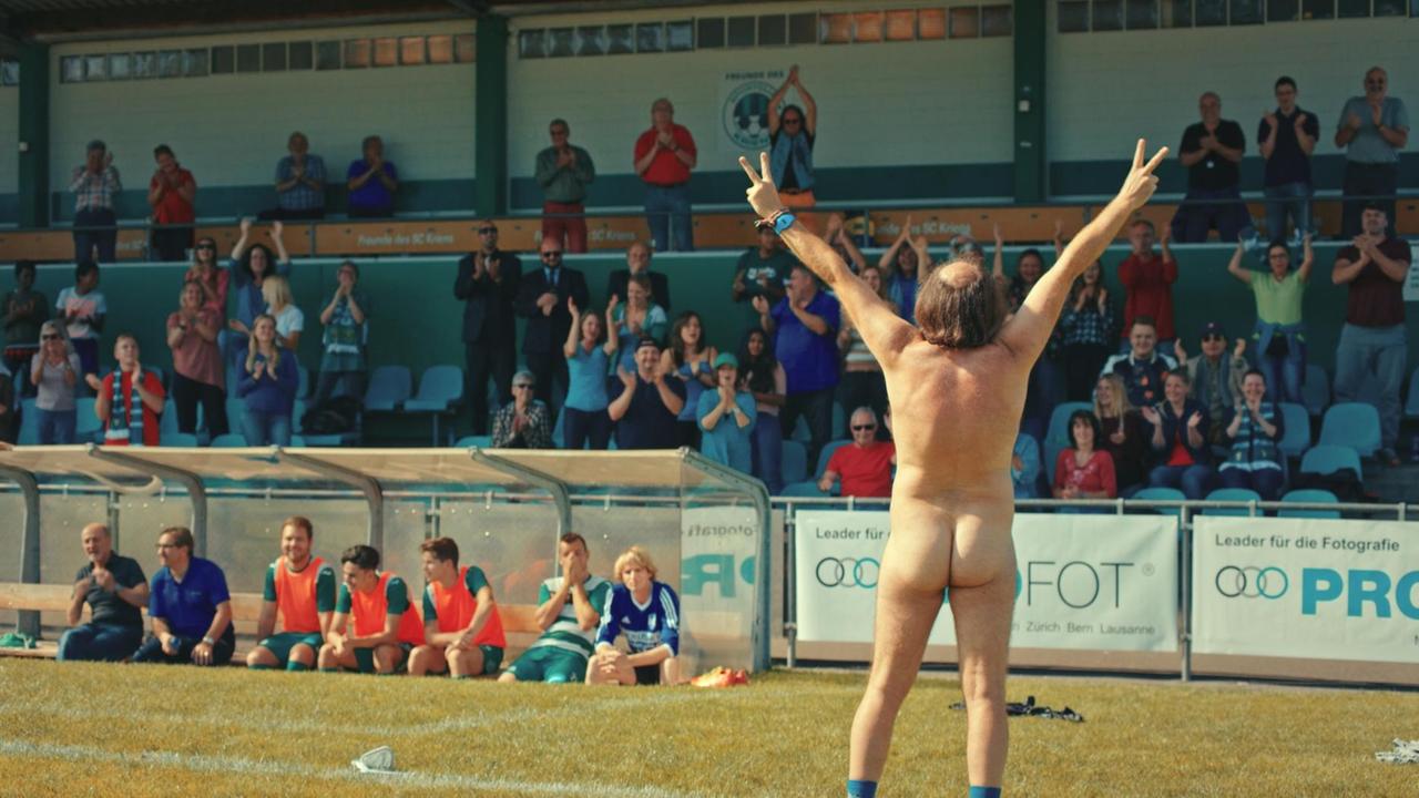 Filmszene aus Flitzer: Ein nackter Mann (Dani Mangisch) steht mit dem Rücken zur Kamera vor dem Stadionpublikum und bildet mit den Fingern ein Siegeszeichen 