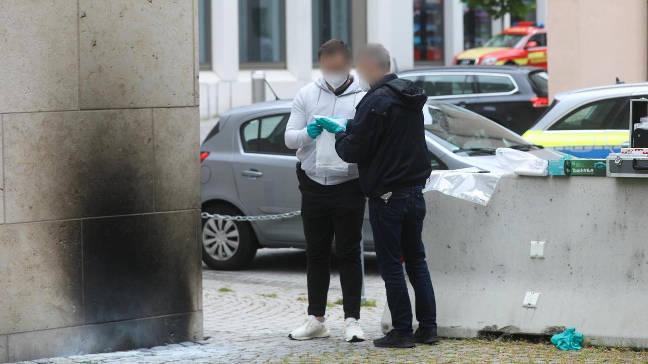 An der hellen Fassade eines Gebäudes ist ein etwa personenhoher Rußfleck zu sehen. Zwei Männer stehen daneben.