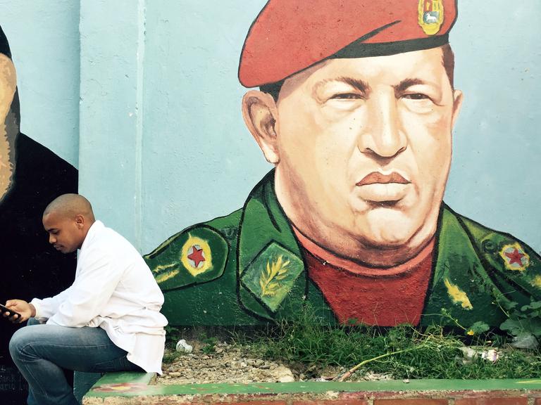 Ein junger Mann sitzt vor einem Graffiti von Hugo Chávez