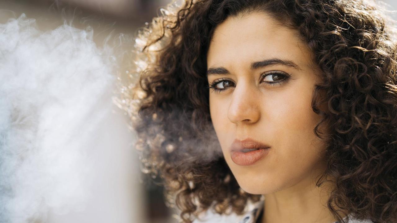 Eine junge Frau mit lockigen braunen Haaren bläst Tabakrauch aus ihrem Mund.