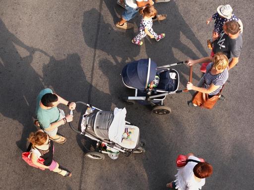 Vogelperspektive: Zwei junge Familien mit Kindern begegenen sich, als sie über eine Straße gehen. Bei der einen Familie schiebt die Mutter den Kinderwagen, bei der anderen der Vater.