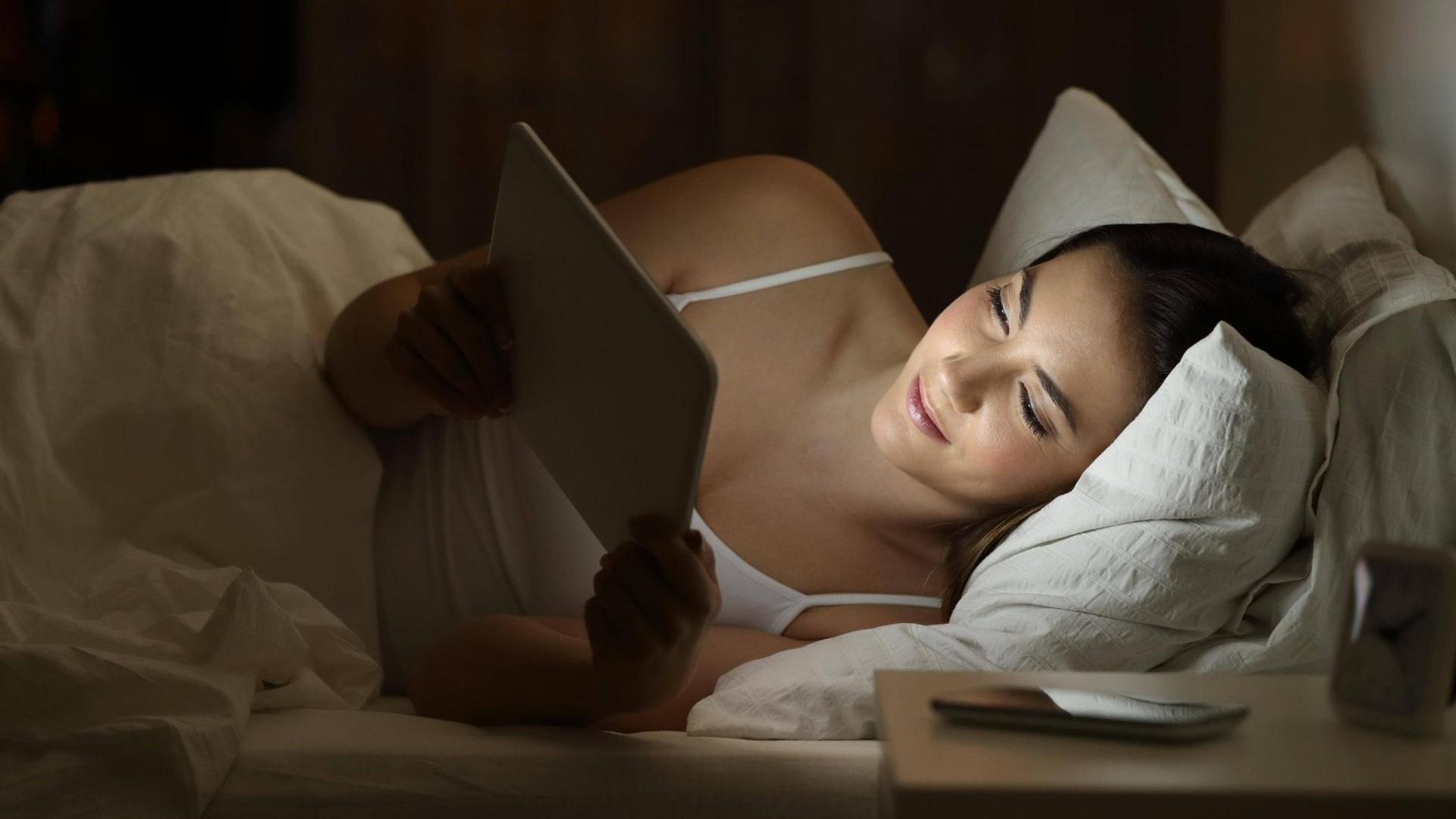 Eine junge Frau schaut nachts in ihrem Bett liegend auf ihr Tablet und lächelt.
