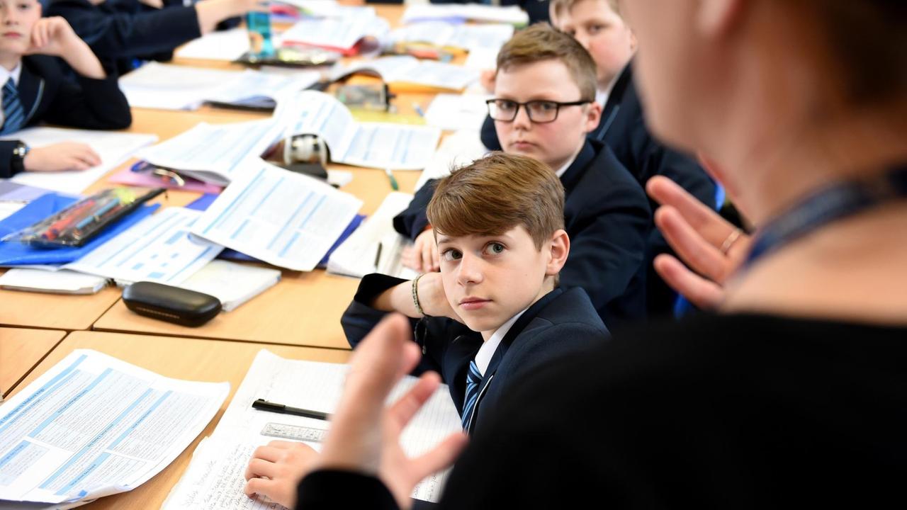 Mehrere jüngere Schüler an Tischen schauen zu einer Lehrerin hoch, die man nur unscharf und im Anschnitt sieht. Sie sagt etwas und gestikuliert mit der rechten Hand.  