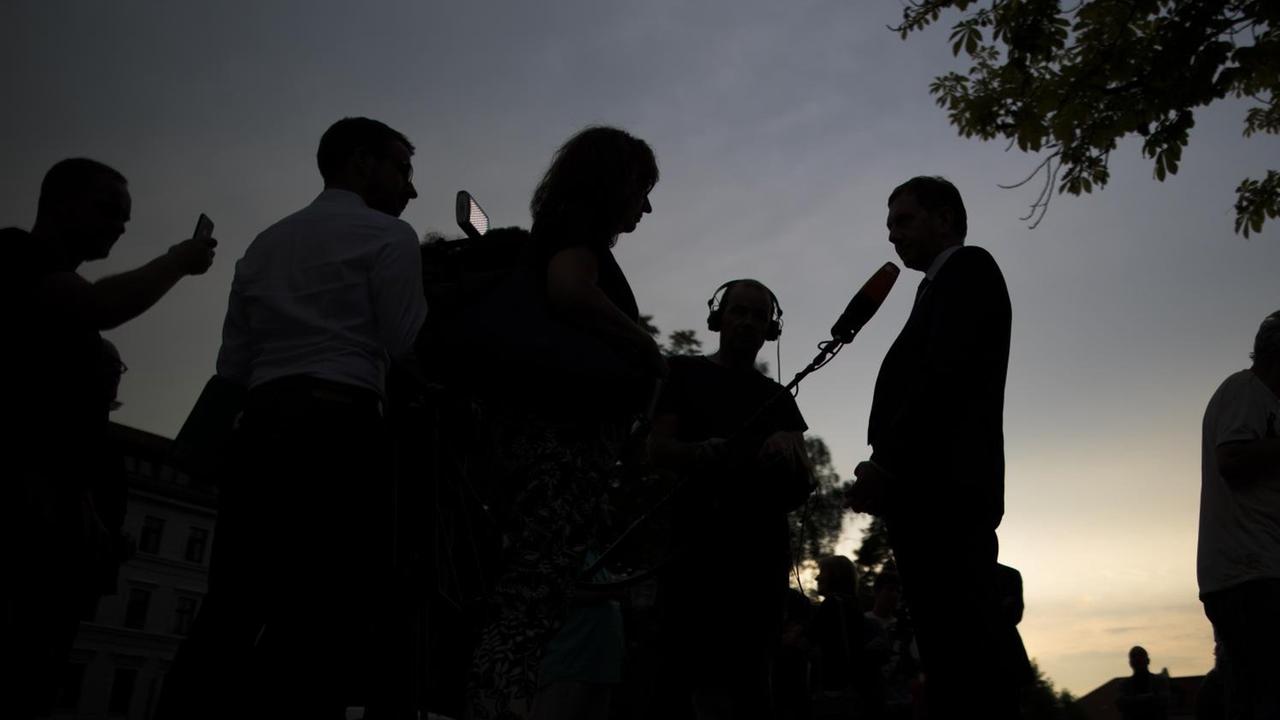 Journalisten, die einen Politiker interviewen, heben sich als schwarze Silhouetten vor einem dämmernden Himmel ab.