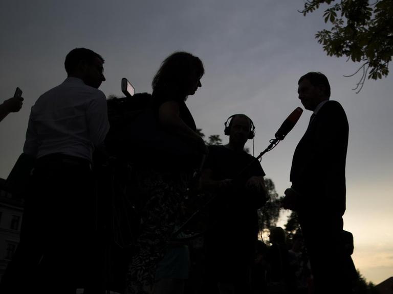 Journalisten, die einen Politiker interviewen, heben sich als schwarze Silhouetten vor einem dämmernden Himmel ab.