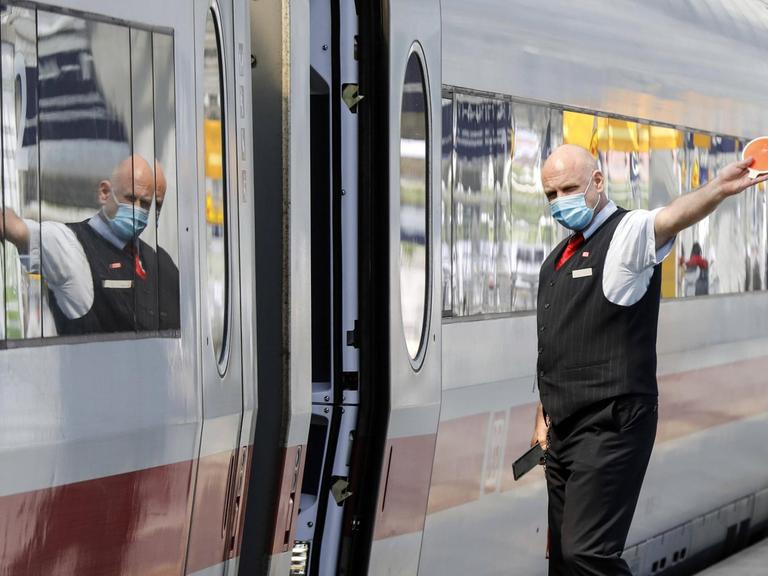 Ein Zugbegleiter mit Gesichtsmaske gibt dem Lokfuehrer das Abfahrtzeichen im Bahnhof Berlin Spandau.