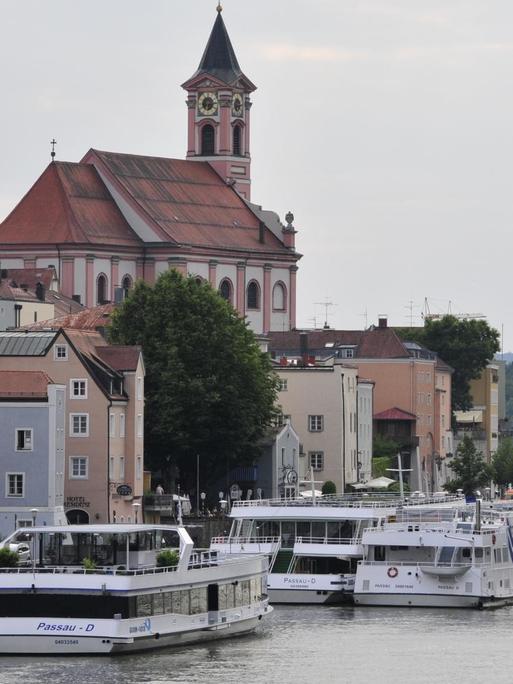 Blick auf die Schiffsanleger und Pfarrkirche St. Paul in Passau. Mehrere Kreuzfahrtschiffe stehen dicht gedrängt am Donauufer.