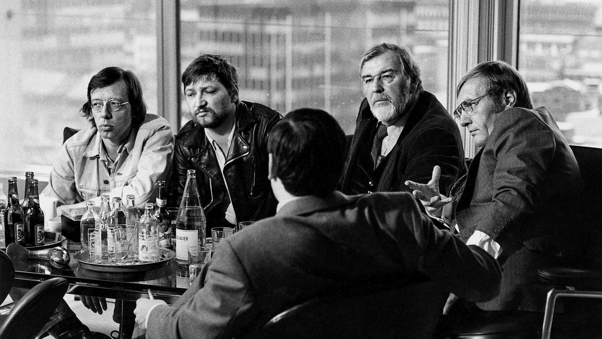 Ein Schwarzweiß-Foto zeigt eine Runde von sechs Männern, an einem Tisch mit Wasser und Bierflaschen sitzend