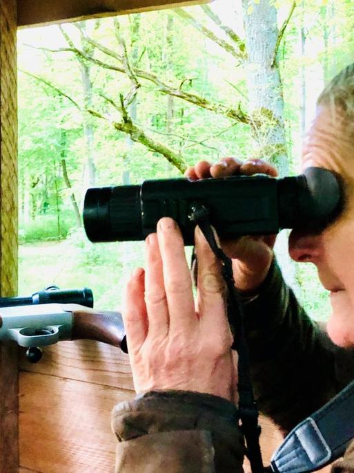 Jägerin Sabine Zuckmantel schaut auf dem Ansitz durchds Fernglas. Im Hintergrund ist ihr Gewehr zu sehen.