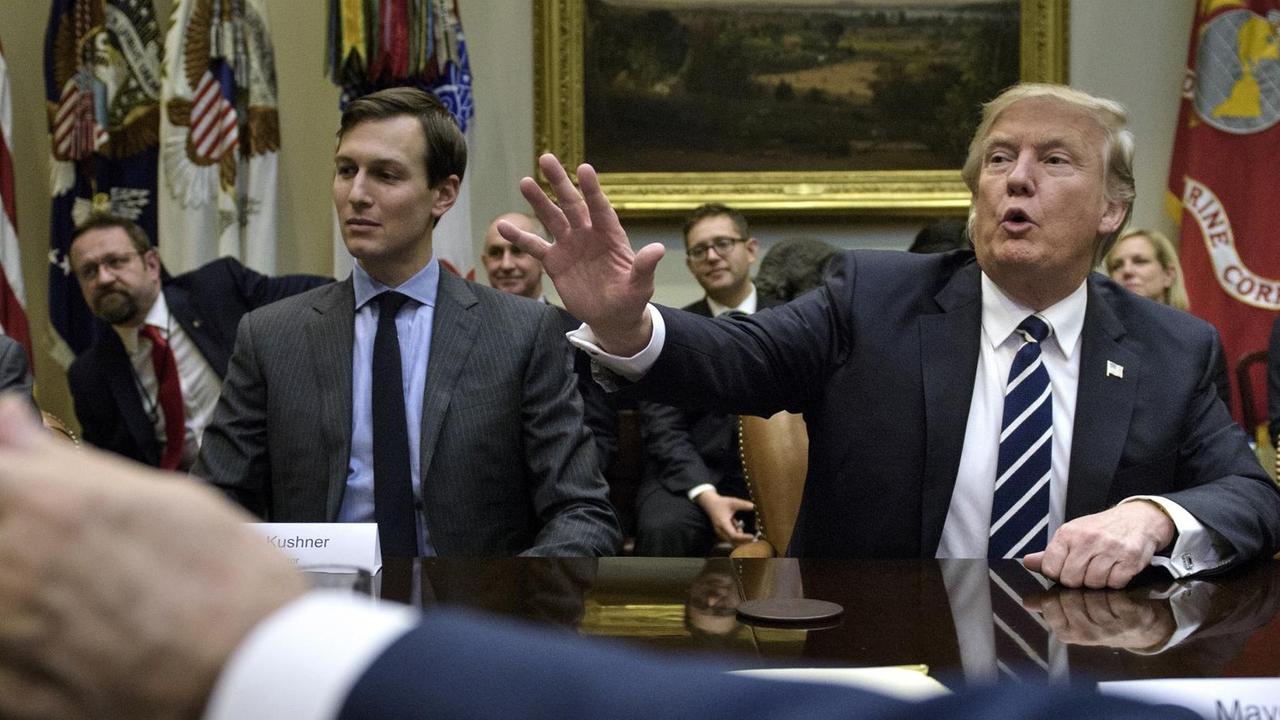 Die drei sitzen nebeneinander, Trump hebt den Arm und sagt etwas. Im Vordergrund unscharf der gestreckte Arm einer anderen Person, im Hintergund weitere Mitarbeiter.