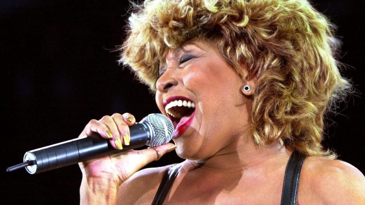 Tina Turner singt in ein Mikrofon. Sie hat kürzere lockige Haare. Und sie hat die Augen zu und lacht beim Singen.