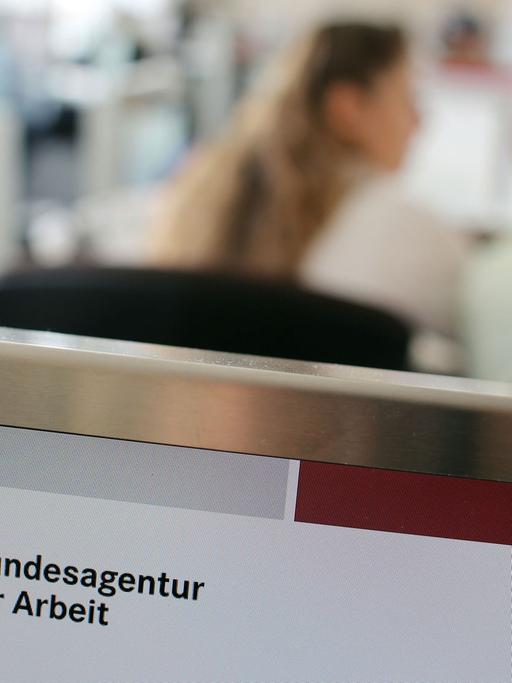 Blick in ein Jobcenter: im Vordergrund das Logo der Bundesagentur für Arbeit, im Hintergrund verschwommen zwei Frauen, die miteinander reden