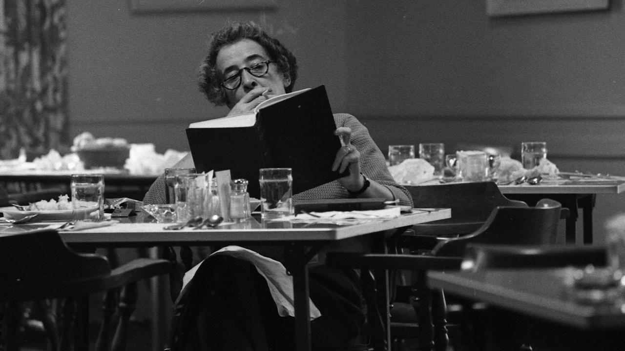 Die Philosophin Hannah Arendt sitzt an einem Tisch in einer Bibliothek, liest und raucht.