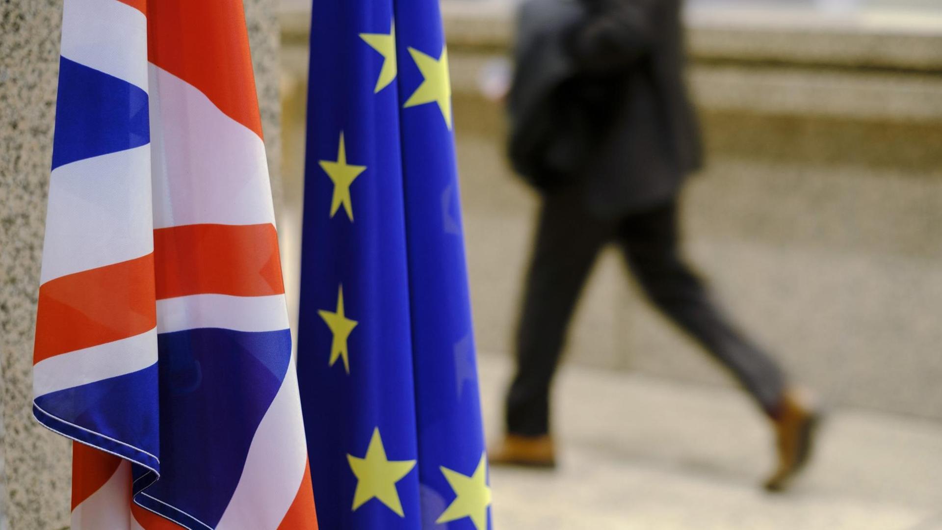 Die Fahnen Großbritanniens und der EU nebeneinander im Vordergrund, im Hintergrund ein laufender Mensch auf der Straße