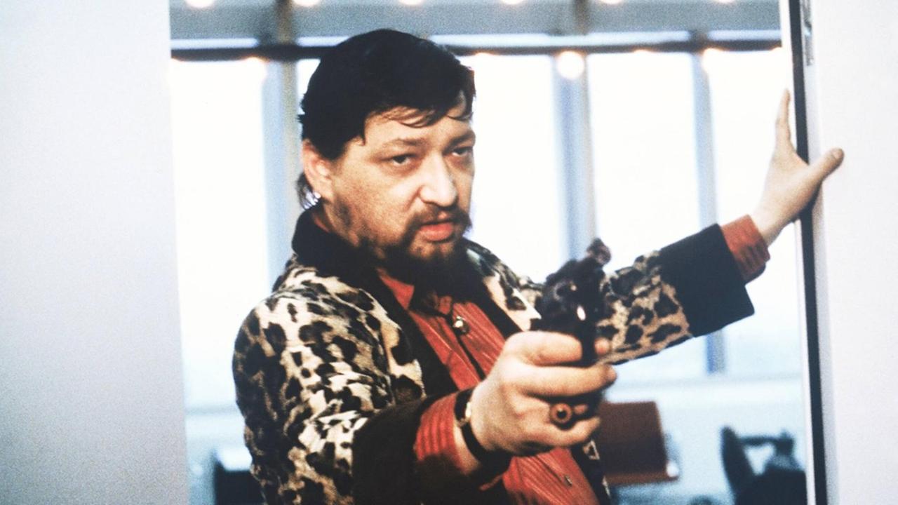Rainer Werner Fassbinder als Schauspieler in einer Szene des Films "Kamikaze 1989". Er hat eine Pistole in der Hand und richtet sie offenbar auf jemand, der nicht zu sehen ist.