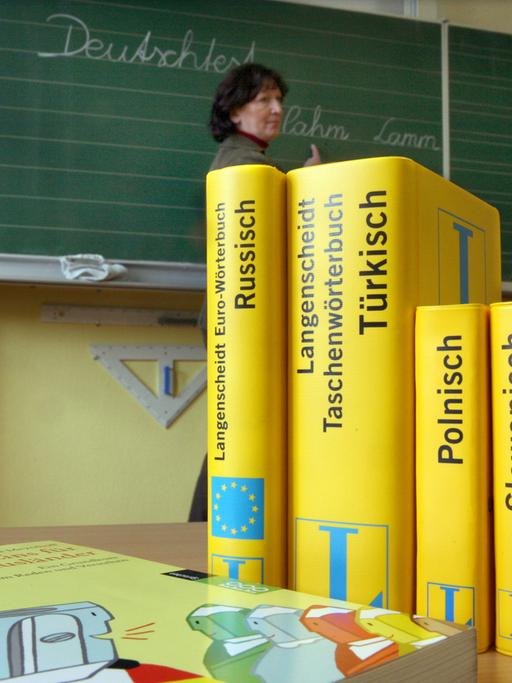 Mehrere Wörterbücher stehen auf einem Tisch in einer Schule. Im Hintergrund ist eine Tafel zu sehen, vor der eine Frau steht.