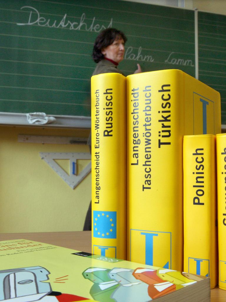 Mehrere Wörterbücher stehen auf einem Tisch in einer Schule. Im Hintergrund ist eine Tafel zu sehen, vor der eine Frau steht.