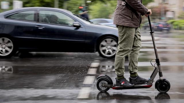 Auf einer nassen Strass fährt im Vordergund ein E-Scooter, im Hintergrund in der Unschärfe sind weitere Autos zu sehen.